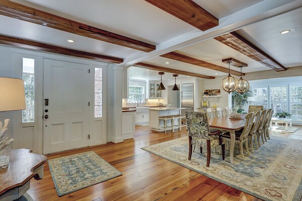 A spacious dining area with a wooden table and chairs, a kitchen in the background, and a large window providing natural light.