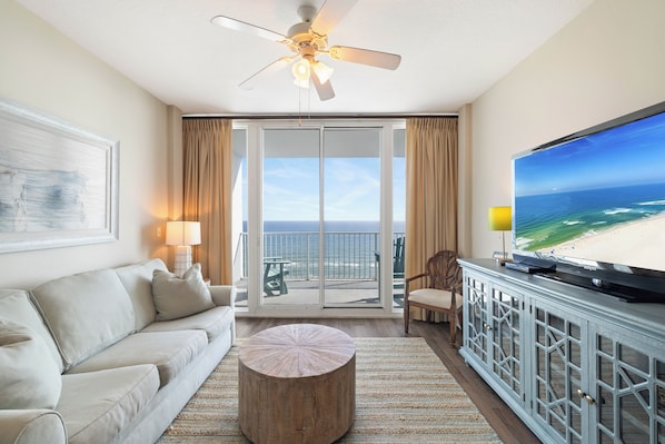 Habitación de hotel con balcón, televisor de pantalla plana grande, sofá, mesa de centro redonda y ventilador de techo.