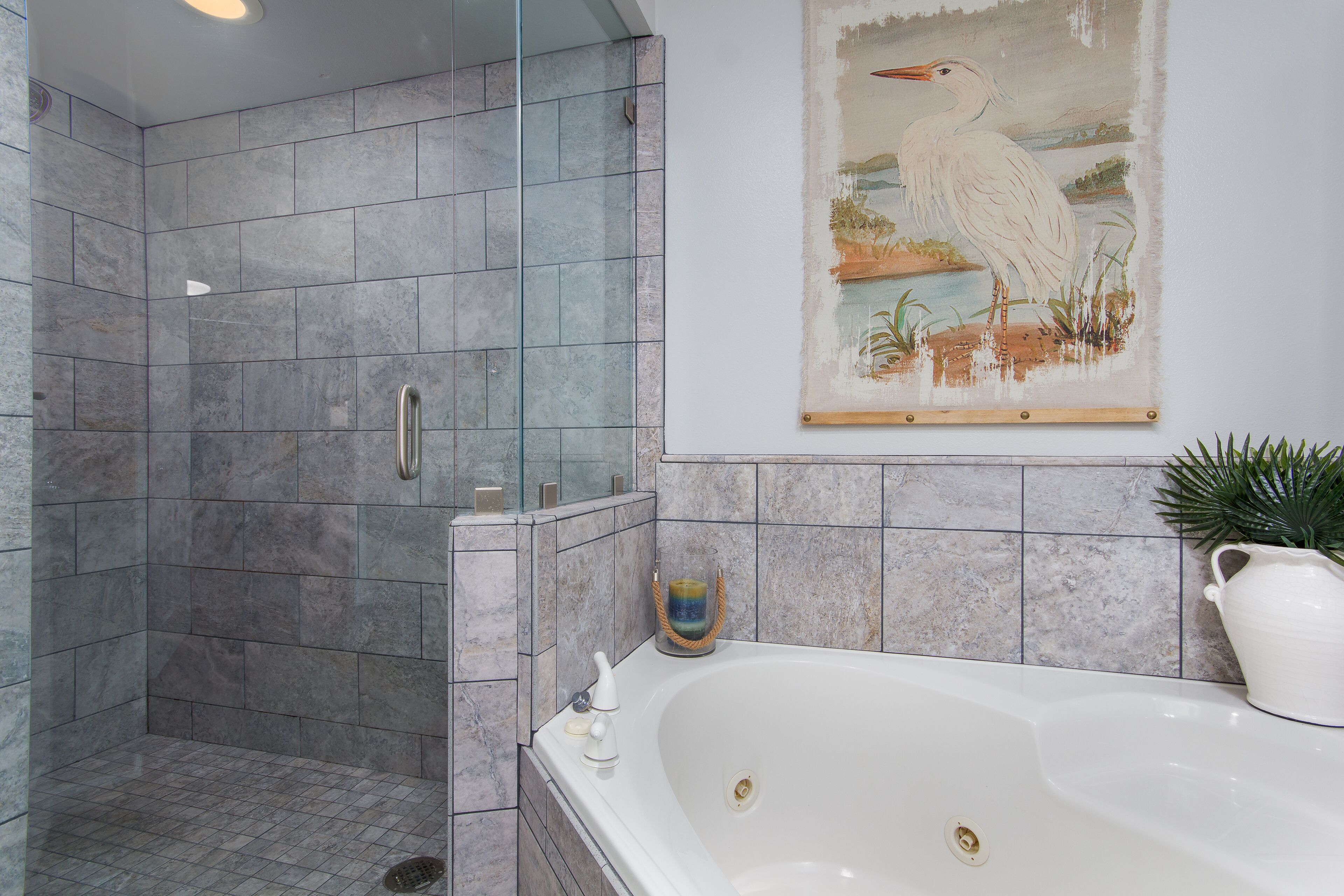 A bathroom with a glass-enclosed shower, a white bathtub, and a painting of a bird on the wall.