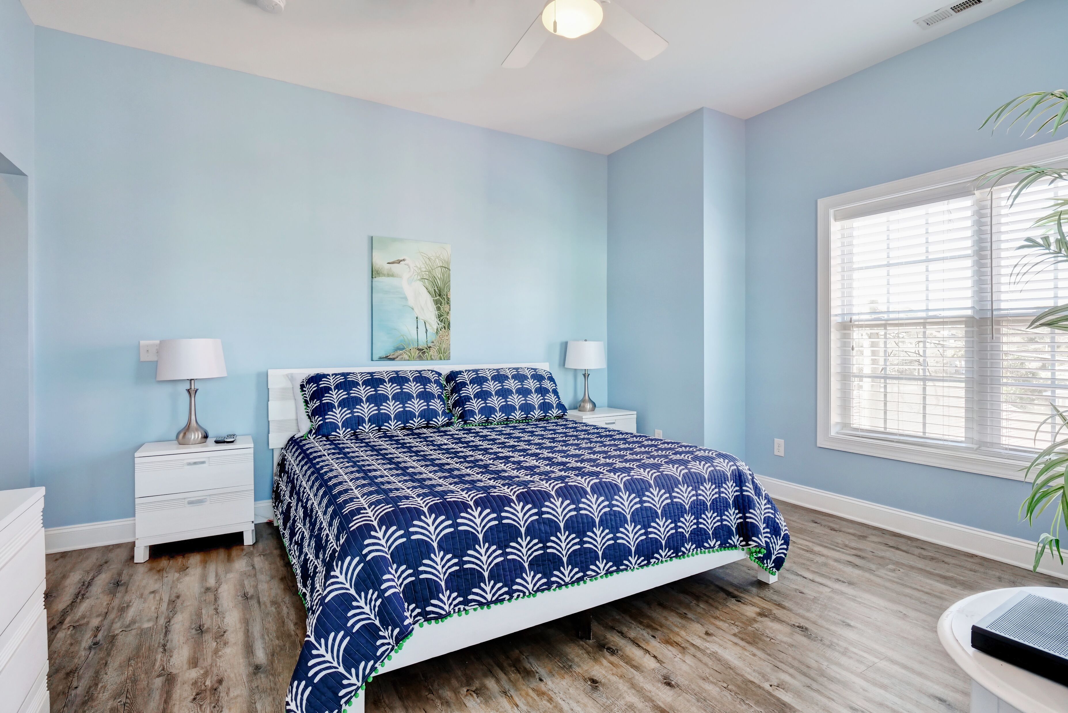 A bedroom with a blue and white patterned bedspread, a nightstand with a lamp, a wall-mounted painting, and a window with blinds.