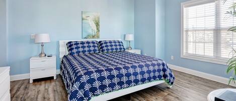 A bedroom with a blue and white patterned bedspread, a nightstand with a lamp, a wall-mounted painting, and a window with blinds.