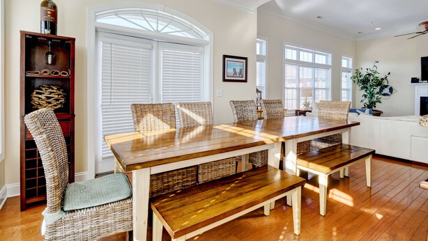 A dining area with a wooden table and chairs, a wicker chair, a wine rack, and a large window with blinds.