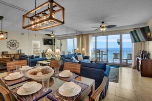 Un vaste espace de vie comprenant un coin repas avec une table dressée, un canapé bleu et un balcon offrant une vue sur l’océan.