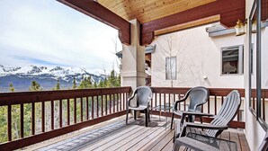 Terraza de madera con sillas y una mesa, con vista a una cadena montañosa nevada.