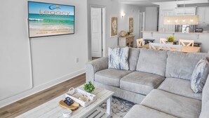 A modern living room with a grey sectional sofa, a white coffee table with a woven basket, and a framed picture of a beach on the wall. - Summer Place #204 - 3 BR Beachfront Condo - Free Beach Service! (Fort Walton Beach)