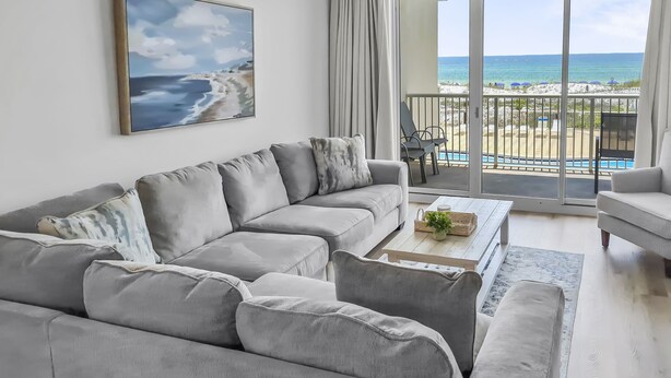 A modern living room with a grey sectional sofa, a coffee table with a plant, and a large window overlooking a beach. - Summer Place #204 - 3 BR Beachfront Condo - Free Beach Service! (Fort Walton Beach)