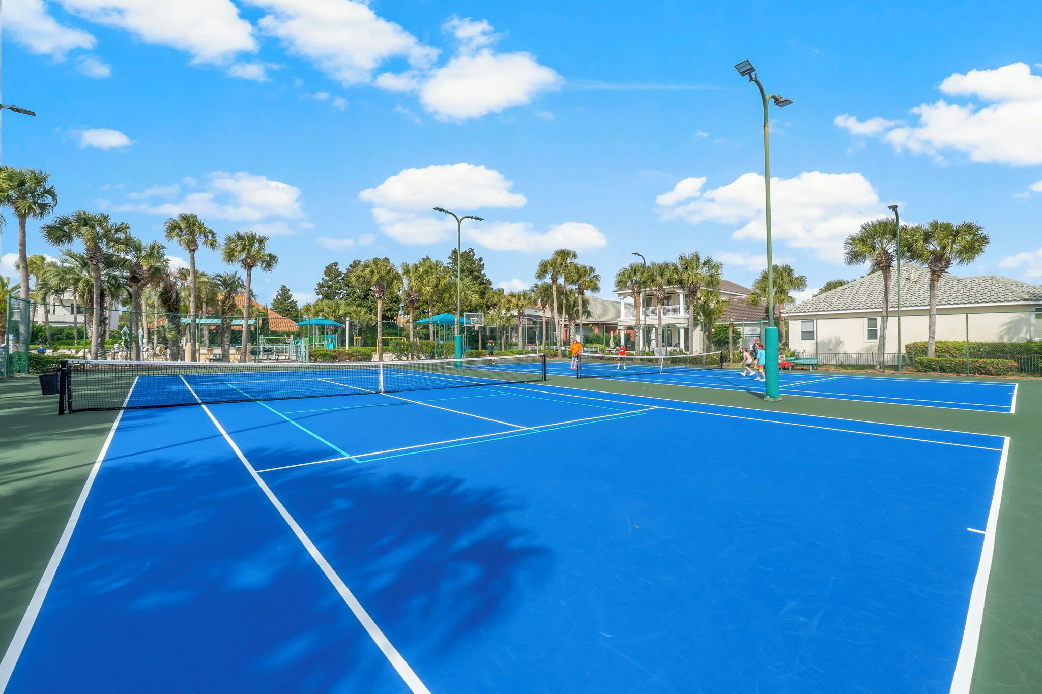 An outdoor tennis court with multiple courts, surrounded by palm trees and buildings.
