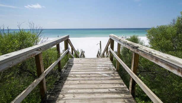 A wooden boardwalk leading to a beach with clear blue water and a sandy shore.