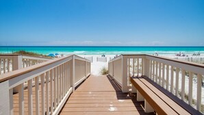 Ein hölzerner Steg führt zu einem Strand mit klarem, blauem Wasser und sandigem Ufer.