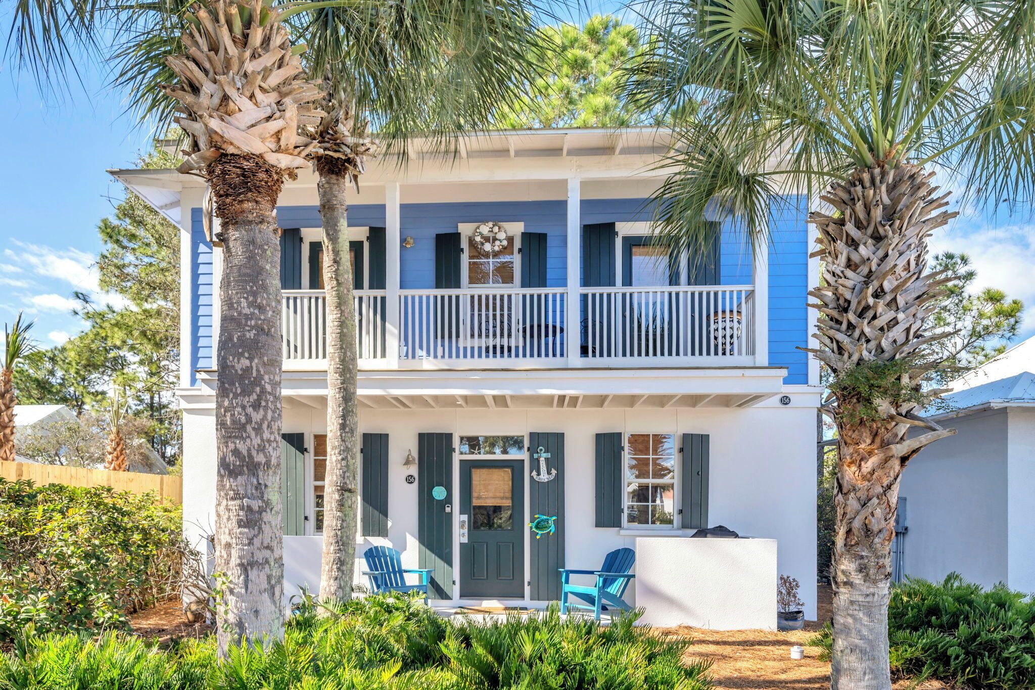 A two-story house with a balcony, a front door with an anchor decoration, and two blue chairs outside.