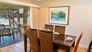A dining area with a wooden table and wicker chairs, a framed picture on the wall, and a view of the outdoors through a glass door. - Ma'alaea Surf Unit E4-2 BR Ground Floor Beachfront Condo, AC, Wi-Fi, Pool, TV (Kihei)