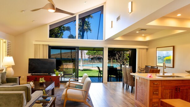 A spacious living area with a large TV, comfortable seating, a kitchen area, and a view of the outdoors. - Ma'alaea Surf Unit E4-2 BR Ground Floor Beachfront Condo, AC, Wi-Fi, Pool, TV (Kihei)
