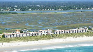 A beachfront hotel with multiple buildings, a sandy beach, and a marshy area in the background. - DR 1309 – Getaway to this oceanfront condo with pool and direct beach access (Wrightsville Beach)