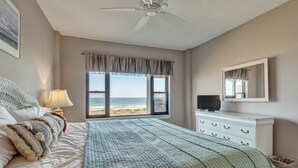 A bedroom with a bed, a dresser, a television, and a large window with a view of the beach. - DR 1309 – Getaway to this oceanfront condo with pool and direct beach access (Wrightsville Beach)