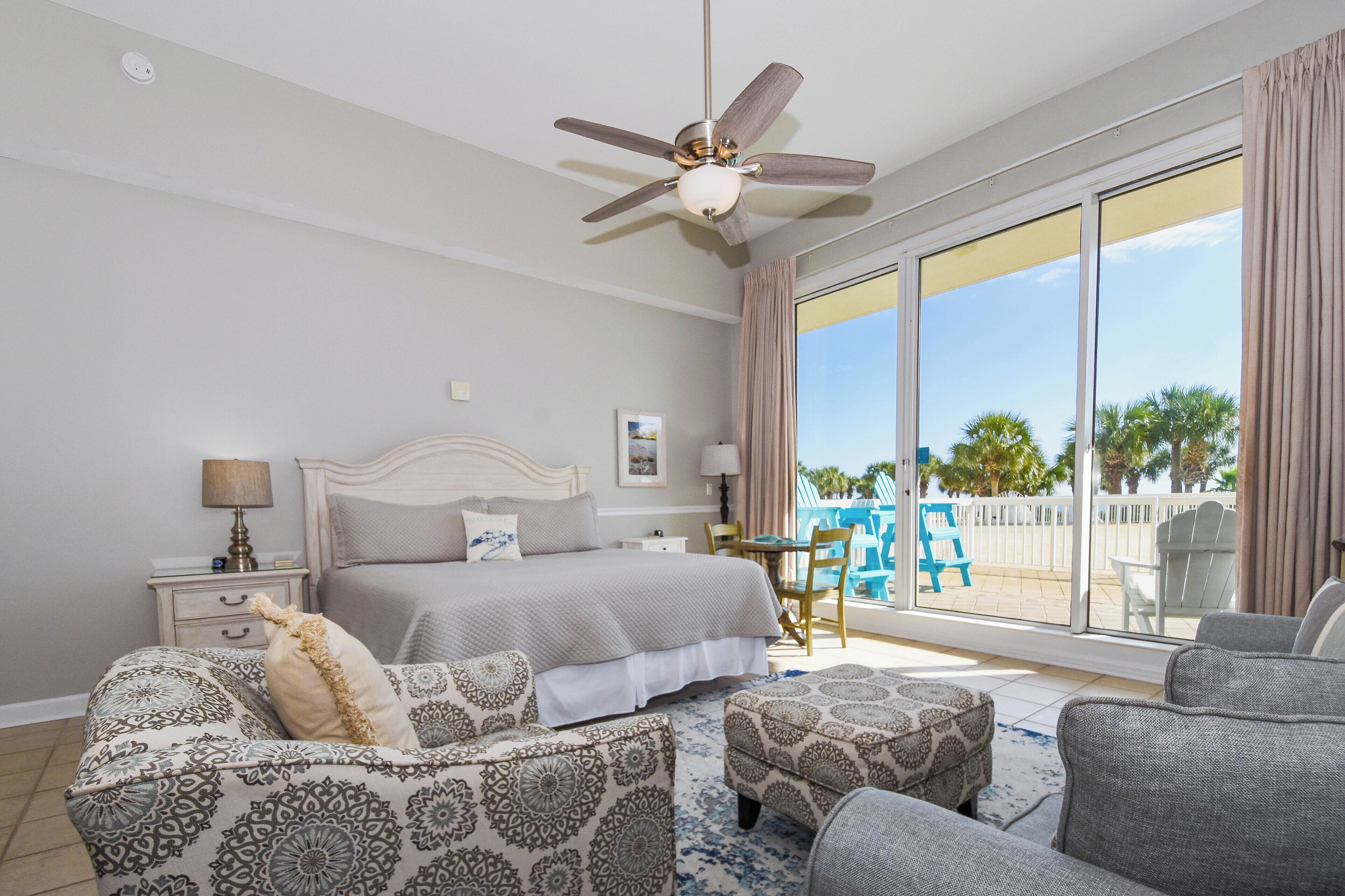 A bedroom with a bed, a ceiling fan, a nightstand, a lamp, a chair, a sofa, a window with a view of palm trees, and a balcony with chairs.