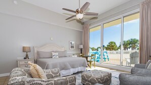 Ein Schlafzimmer mit einem Bett, Deckenventilator, Nachttisch, Lampe, Stuhl, Sofa, Fenster mit Blick auf Palmen und einem Balkon mit Sitzgelegenheiten.