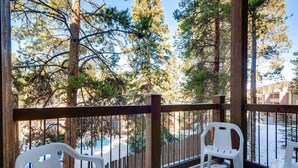 A wooden deck with two white plastic chairs, overlooking a snowy landscape with trees and a building in the background. - Adorable Condo, Walking Distance To Everything. Including Pool/Hot Tub IC4 (Breckenridge)