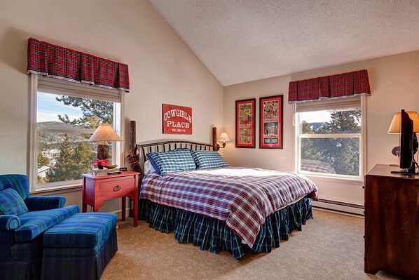 A bedroom with a bed, a nightstand, a chair, a desk, and two windows with plaid curtains.