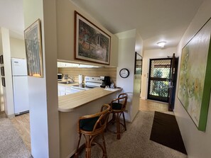 Een compacte keuken met een magnetron, fornuis en koelkast. Er staan twee rieten barstoelen en er hangt een schilderij boven de toonbank.