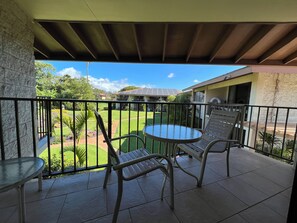 A balcony with a glass table and two chairs, overlooking a well-maintained garden and a house with solar panels.