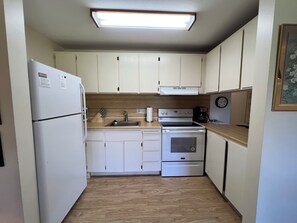 A compact kitchen with white cabinets, a refrigerator, a sink, an oven, and a clock on the wall.
