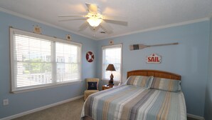 A bedroom with a bed, a ceiling fan, two windows with blinds, a chair, and a sail sign on the wall.