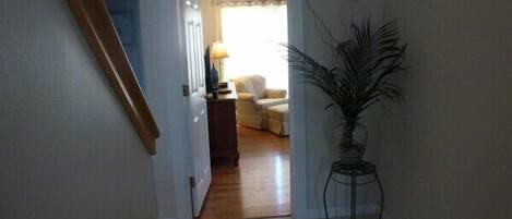 A hallway with a wooden staircase, a door leading to a living room with a sofa and a lamp, and a potted plant.