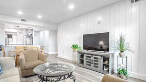 A modern living room with a glass coffee table, beige sofa, and a flat-screen TV on a wooden cabinet. - Shorewood 332 - Oceanview 3rd Floor Condo (Hilton Head Island)