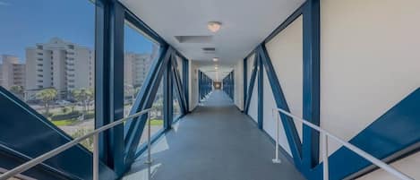 Een gang met grote ramen, blauwe structurele balken en een helder uitzicht op gebouwen.
