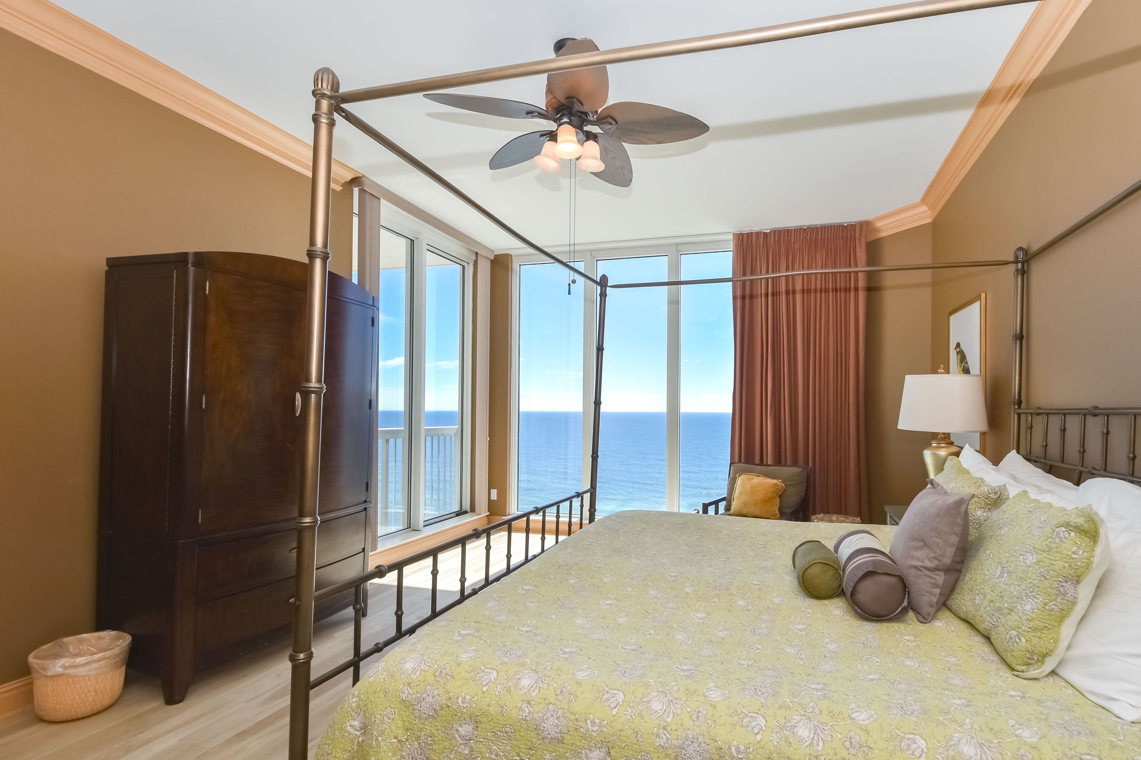 Une chambre avec balcon, un lit recouvert d’une literie à motifs floraux, une armoire en bois et un ventilateur de plafond.