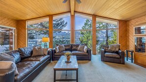 A cozy living room with a leather sofa, a wooden coffee table, and a ceiling fan. Large windows offer a view of trees and mountains.