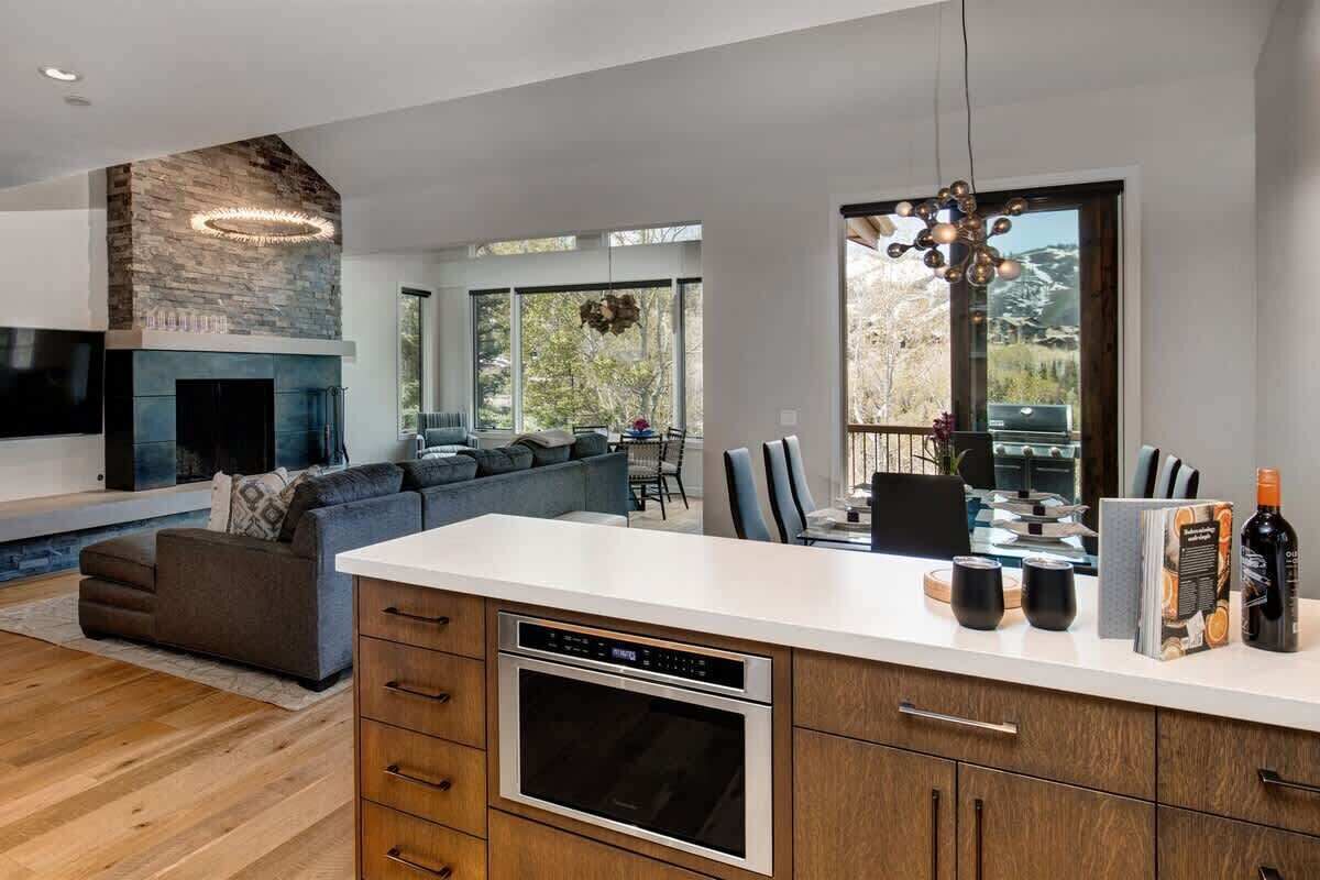 A modern kitchen with a built-in oven, a dining area with a table and chairs, a fireplace, and large windows offering a view of the outdoors.