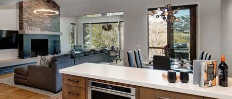 A modern kitchen with a built-in oven, a dining area with a table and chairs, a fireplace, and large windows offering a view of the outdoors.