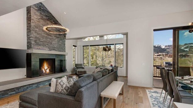 Uma sala de estar moderna com lareira, sofá seccional, um banquinho e área de jantar com vista para as montanhas.