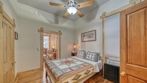 Une chambre avec un plancher en bois, un lit recouvert d’une courtepointe, un ventilateur de plafond, un radiateur et une porte.