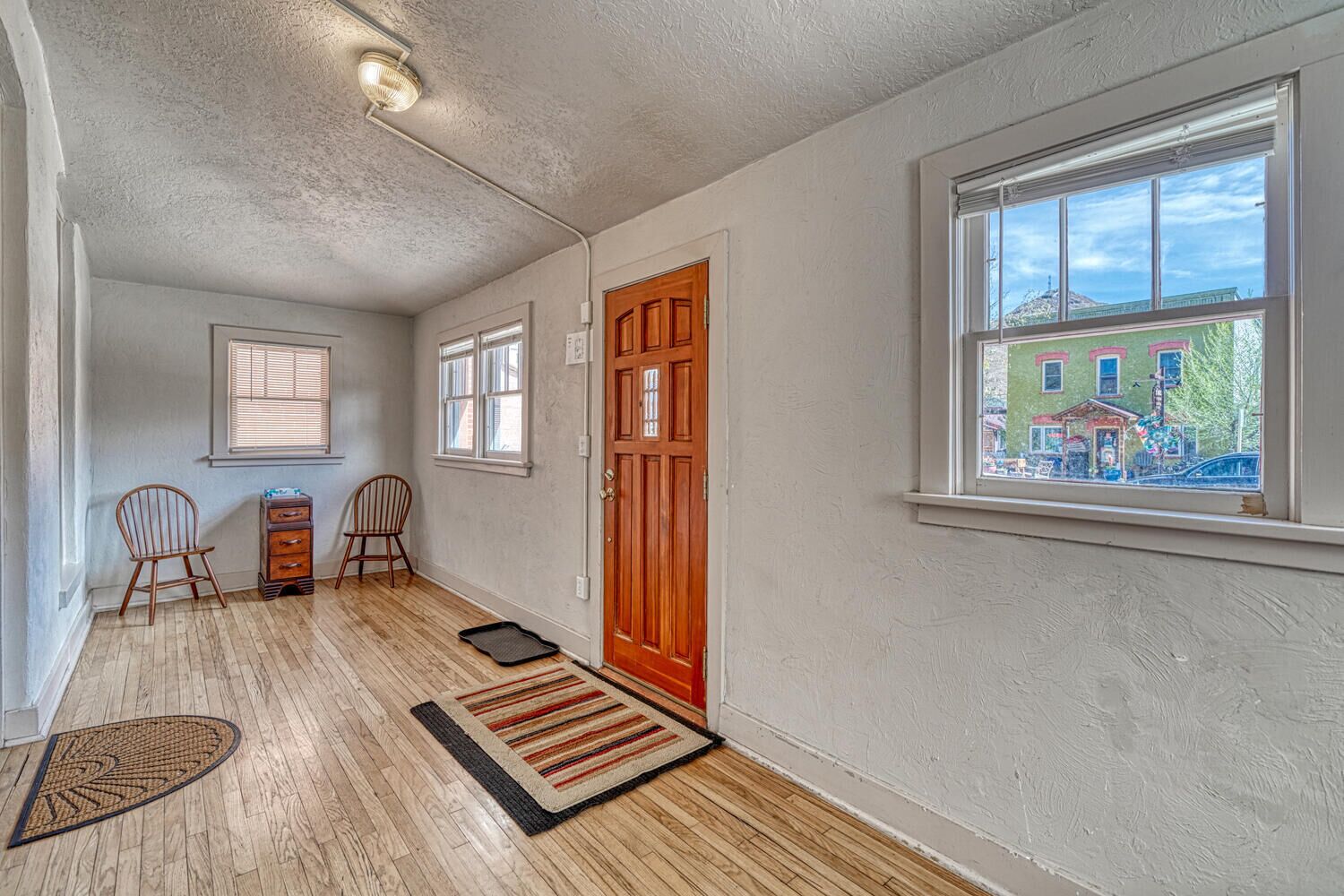 A hallway with wooden flooring, a door, two chairs, a small table, and a window with a view of buildings.