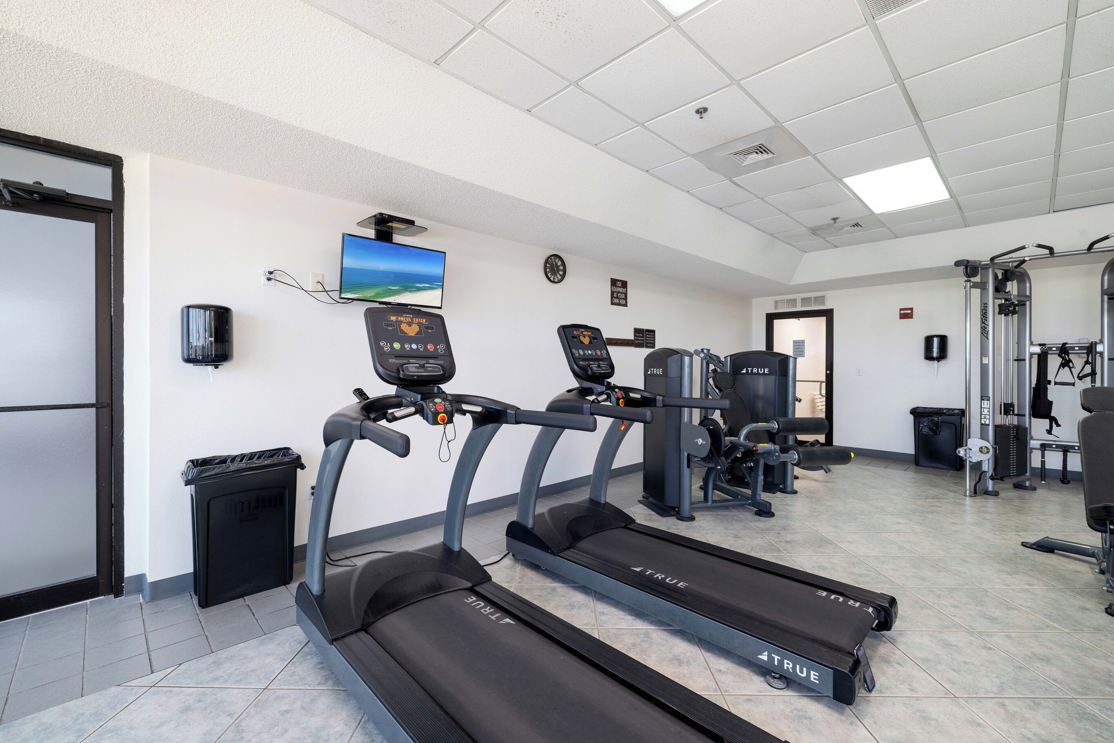 Ein Fitnessstudio mit Laufbändern, Kraftgeräten und einem Fernseher an der Wand.