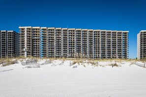 A beachfront high-rise building with multiple balconies, a sandy foreground, and a clear blue sky. - Phoenix VII 1206 | Heated Pool | Tennis | My Beach Getaways (Orange Beach)
