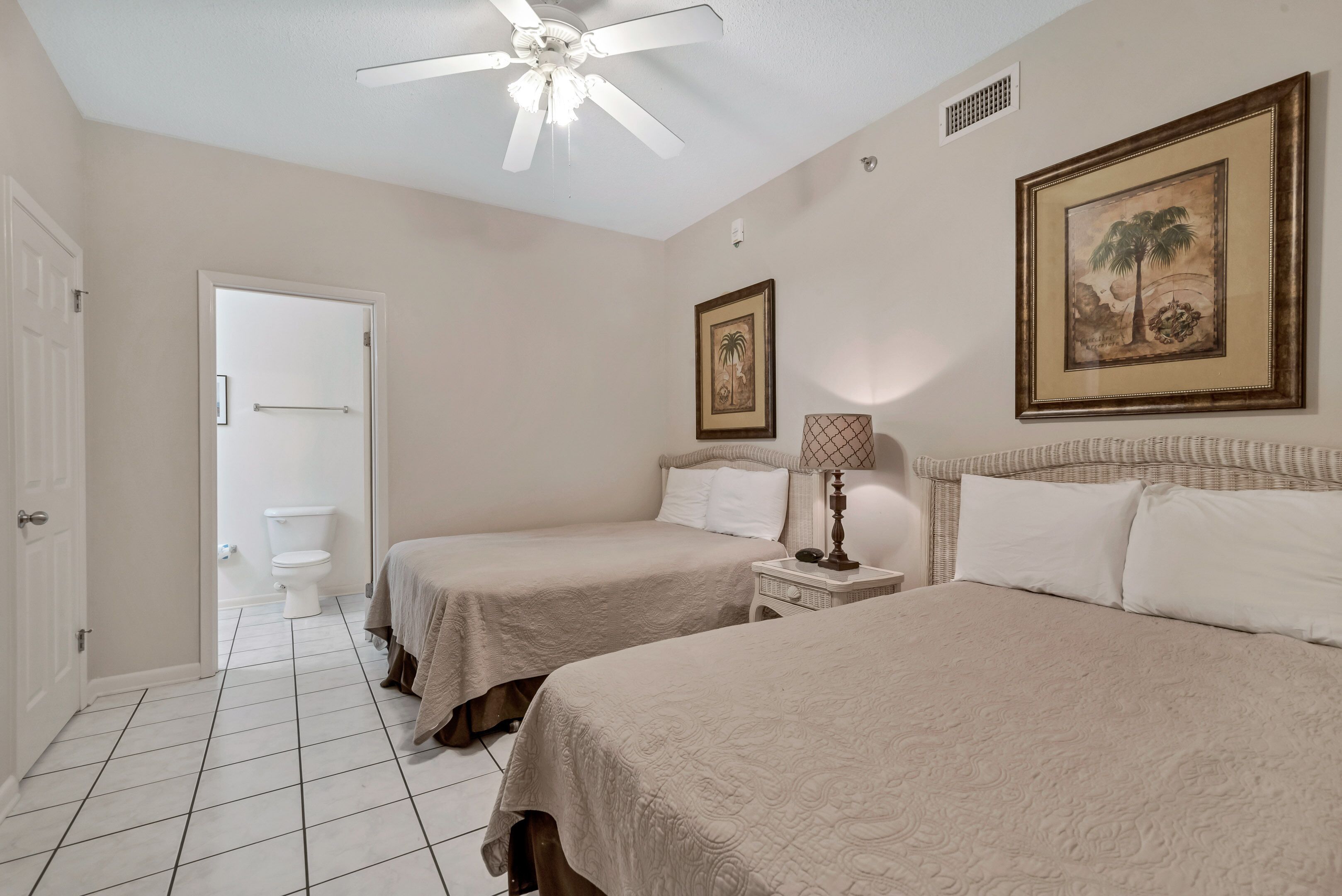 A hotel room with two beds, a ceiling fan, a framed picture, and a bedside table with a lamp.