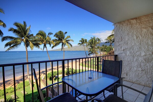 A balcony with a table and chairs, overlooking a beach with palm trees and the ocean.