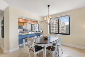 A modern kitchen with a central island, a dining area with a round table and chairs, and a large window offering a view of buildings.
