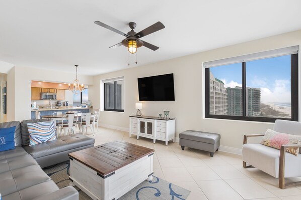 A modern living room with a sofa, coffee table, and a television. There is a kitchen in the background with a dining area.