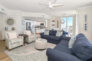 Un salon spacieux avec un canapé bleu foncé, un fauteuil blanc et un pouf assorti. On peut voir un ventilateur de plafond et un lustre.