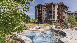 Un jacuzzi rodeado de rocas y árboles, con un edificio de varios pisos al fondo.