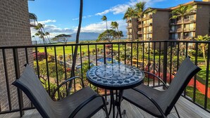 A balcony with a round table and two chairs, overlooking a pool and lush greenery. - Kauhale Makai #330 Oceanfront, Pool, Hot tub, A/C, Wi-Fi, Stunning Sunsets! (Kihei)