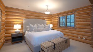 Quarto de cabana de toras com uma cama, duas mesas de cabeceira, um abajur, uma janela e uma caixa de armazenamento trançada.