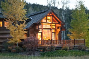 Une cabane en rondins dotée d’une grande véranda et de fenêtres, entourée d’arbres et d’une zone boisée.