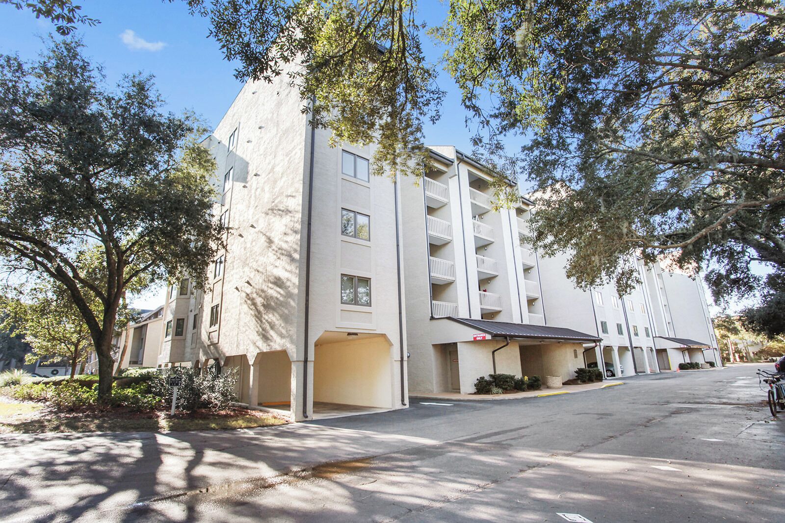 Un edificio residencial de varios pisos con balcones, un área de estacionamiento techada y árboles en primer plano.