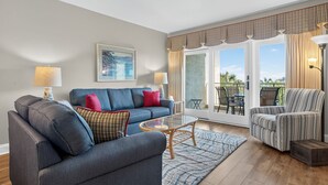 Ein Wohnzimmer mit einer Couch, einem Sessel und einem Glastisch. Ein großes Fenster mit Vorhängen bietet Blick ins Freie.