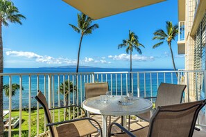 Ein Balkon mit rundem Tisch und vier Stühlen, mit Blick auf ein klares blaues Meer, Palmen und eine entfernte Bergkette.